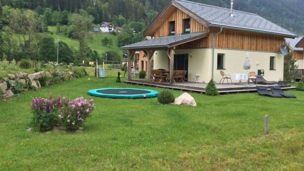 A wooden house with a porch, a trampoline, and a small garden with purple flowers.