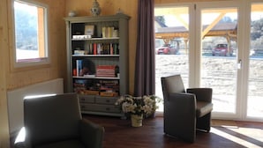 A cozy room with a wooden interior, a bookshelf, two armchairs, and a view of a snowy landscape through large windows.