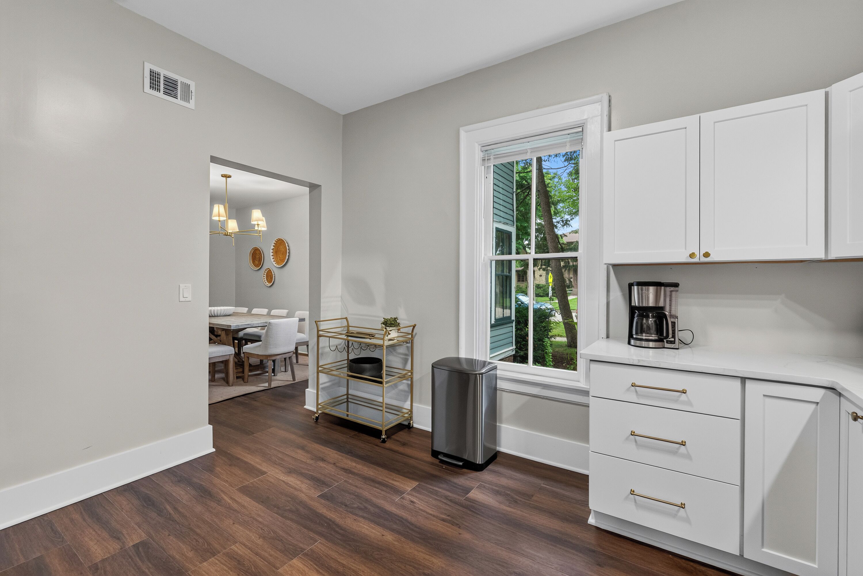 A modern kitchen with white cabinetry, a coffee maker, and a dining area with a table and chairs.