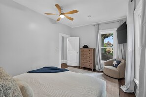 Ein Schlafzimmer mit einem Bett, einem Schminktisch, einem Sessel, einem Deckenventilator und einem Fenster mit Vorhängen.