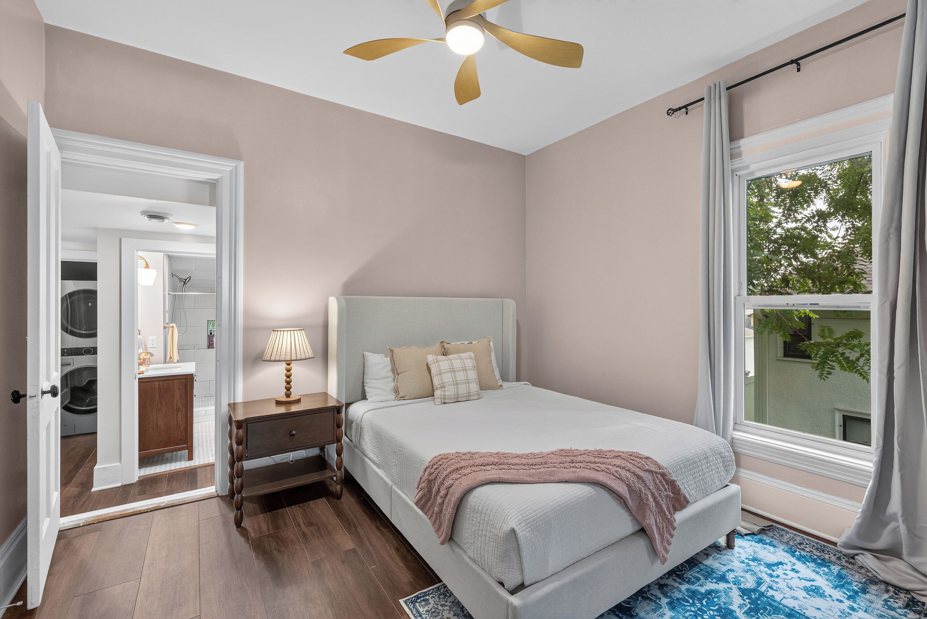A bedroom with a bed, a nightstand, a lamp, a ceiling fan, and a window with a view of greenery.
