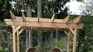 A wooden pergola with two wicker swing chairs and a black table in a garden setting. - Victorian · Game Room · Walk to Beach + Downtown (Lake Geneva)