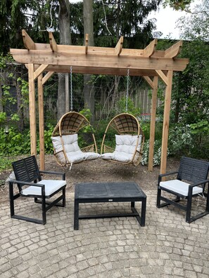 Puinen pergola, jossa on kaksi rottinkista riipputuolia ja musta pöytä, puutarhamaisessa ympäristössä.