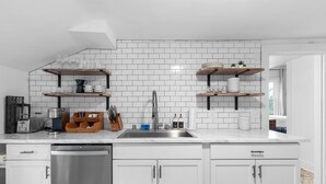 A modern kitchen with white cabinets, a stainless steel dishwasher, and a marble countertop. There are two wooden shelves above the sink, holding glassware and kitchen items. - Victorian · Game Room · Walk to Beach + Downtown (Lake Geneva)