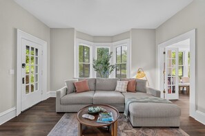 Un salon comprenant un canapé d’angle, une table basse et une lampe. De grandes fenêtres équipées de stores, une porte et une vue sur la verdure à l’extérieur complètent la pièce.