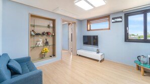 Uma sala de estar moderna com um sofá azul, uma estante de vidro com objetos decorativos, um console branco, uma televisão de tela plana e uma janela com vista.