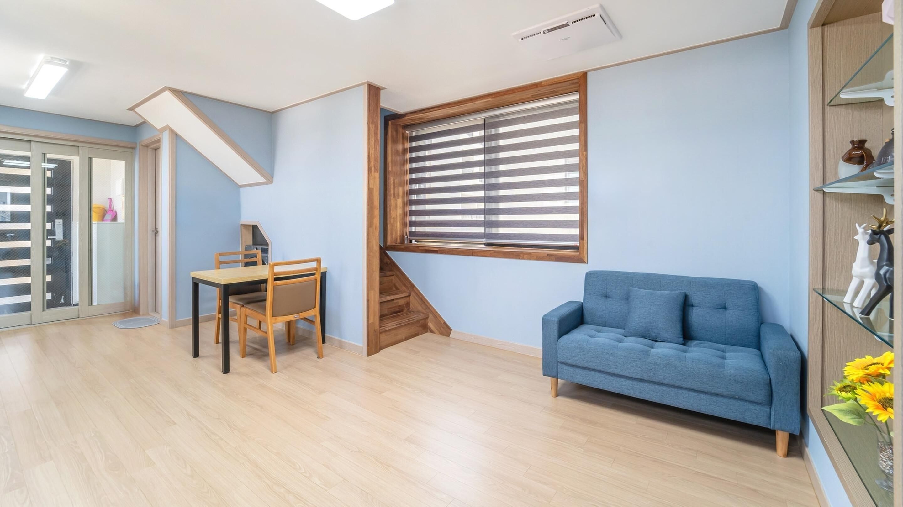 Una habitación con un sofá azul, una mesa de comedor de madera con sillas, una escalera y una ventana con persianas.