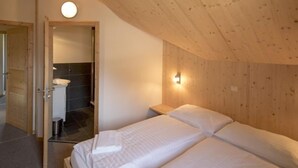 Quarto com duas camas, teto de madeira, luminária redonda e banheiro visível por uma porta aberta.
