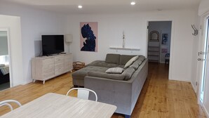 Un salon moderne avec un canapé gris, un téléviseur à écran plat posé sur un meuble en bois, une table à manger avec des chaises et un plancher en bois.