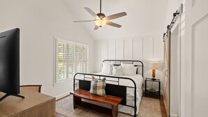 A bedroom with a bed, a ceiling fan, a bedside table with a lamp, a bench, and a TV on a wooden stand. - Stunning backyard oasis nestled between pier/village and beach - SSI Sunshine (St Simons Island)