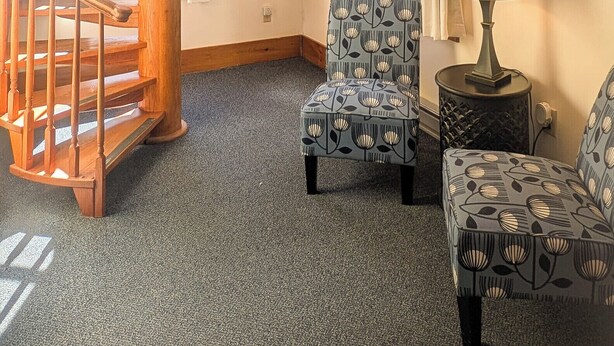 Una habitación con una escalera espiral de madera, dos sillones estampados, una mesita con lámpara y un ventanal con cortinas.