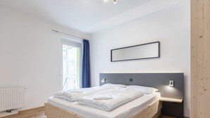 A modern bedroom with a wooden bed, a large window with curtains, a white radiator, and a wall-mounted light fixture. - für bis zu 8 Personen mit IR Sauna by Interhome (Eisenerz)