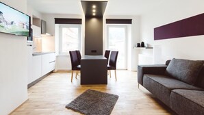 A modern living room with a sofa, a TV mounted on the wall, a dining table with chairs, and a kitchen area with cabinets and a window.