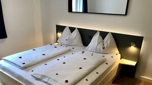 Un letto ben rifatto con lenzuola bianche e petali di rosa, un comodino con una lampada e uno specchio a parete.