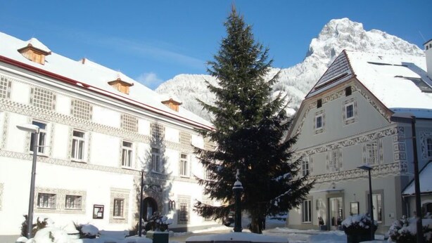 Un cortile innevato con un grande abete, una panchina e edifici dall'architettura tradizionale.