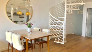 Ein Essbereich mit einem runden Spiegel, einem Holztisch und Stühlen mit flauschigen Kissen. Im Hintergrund ist eine Wendeltreppe zu sehen.
