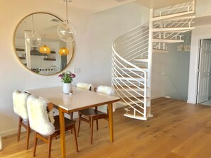 A dining area with a round mirror, wooden table, and chairs with fluffy cushions. A spiral staircase is visible in the background.