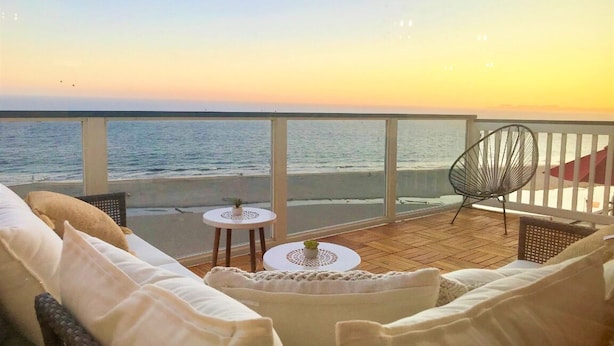 Ein Balkon mit Meerblick, eine Rattan-Couch, zwei kleine runde Tische und ein Metallstuhl.
