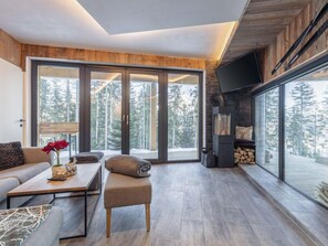 A modern living room with a sofa, coffee table, and ottoman, large sliding glass doors leading to a forest view, and a fireplace.