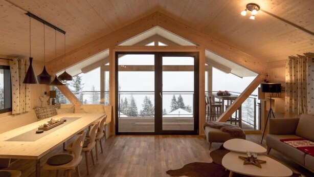 Interior acolhedor de uma cabana com área de jantar, sala de estar e varanda com vista para árvores cobertas de neve.