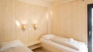 Quarto de madeira com duas camas, cada uma com roupa de cama e toalhas brancas. Há luzes fixadas na parede e uma porta visível.