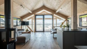 Um amplo loft com teto alto, lareira a lenha embutida, ampla porta de vidro deslizante com vista para as montanhas e área de cozinha minimalista.