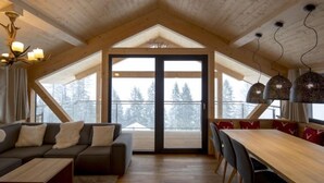 A cozy interior with a wooden ceiling, a sofa, a dining table with chairs, and a view of snow-covered trees through large windows. - Premium # 01 IR-Sauna&Sprudelpool aussen by Interhome (Pichl)