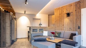 A modern living room with a grey sofa, a wooden coffee table, and a bench. There is a kitchen area in the background with wooden walls and a hanging light fixture. - Superior # 03 Sauna&Sprudelwanne innen by Interhome (Pichl)