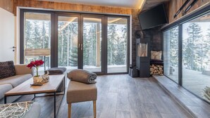 Ein modernes Wohnzimmer mit Sofa, Couchtisch und Hocker, großen Schiebegläsern mit Blick auf einen Wald und einem Kamin.