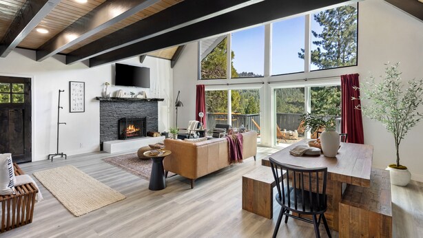 A spacious living room with a fireplace, wooden beams, large windows, and a dining area with a table and chairs.