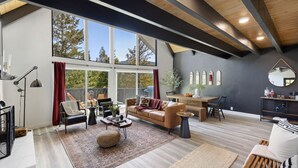 A modern living room with a large window, a brown leather sofa, a coffee table, and a dining area with a wooden table and chairs.