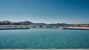 A large, clear swimming pool with umbrellas and lounge chairs, surrounded by a beach and mountains. - Cosme, A Luxury Collection Resort, Paros (Paros)