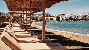 A beachside seating area with sun loungers and umbrellas, overlooking a calm sea and a coastal town. - Cosme, A Luxury Collection Resort, Paros (Paros)