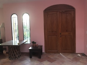 A room with pink walls, a wooden door, a window with metal bars, a small table with a potted plant, and a tiled floor.