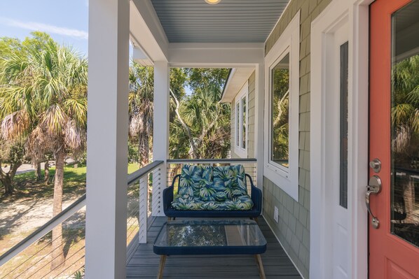 A covered porch with a single blue chair and a glass-top table, surrounded by palm trees and a red door.