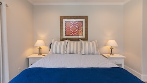 Een slaapkamer met een bed, twee nachtkastjes, een lamp op elk en een lijst met een kunstwerk erboven, plus een raam met gordijnen.