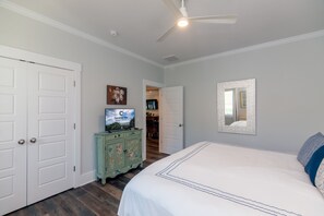 Une chambre à coucher avec un lit, un téléviseur posé sur un meuble, un ventilateur de plafond et une porte donnant sur une autre pièce.