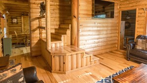 Ein Holzblockhaus-Interieur mit einer Holztreppe, einem Kamin, einem Ledersofa und einem Holzessttisch.