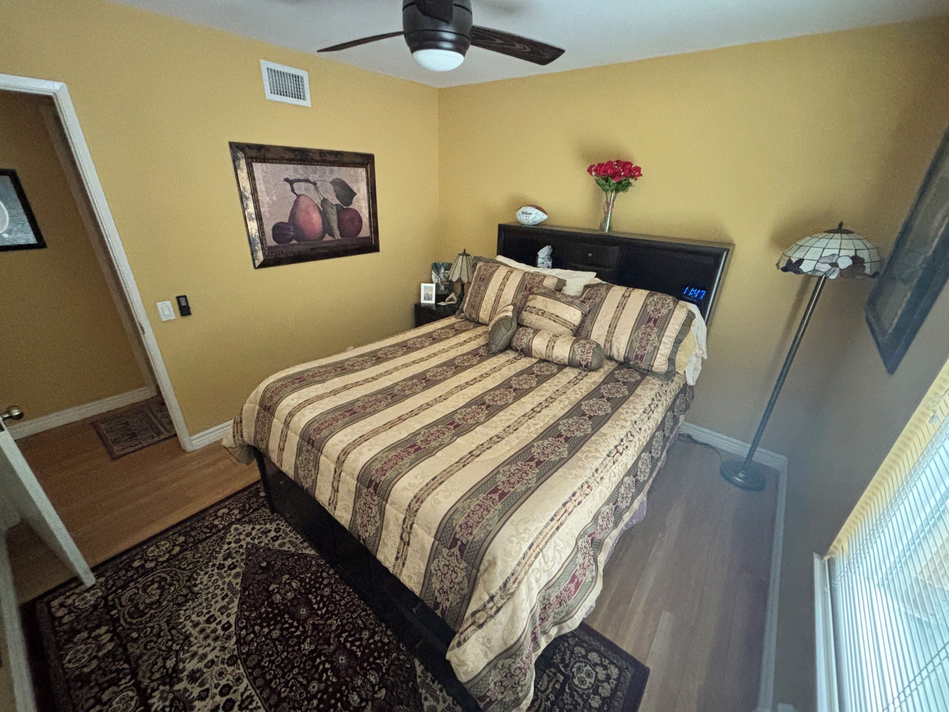 Une chambre à coucher avec un lit, une table de chevet, une lampe, un tableau au mur et une fenêtre avec des stores.