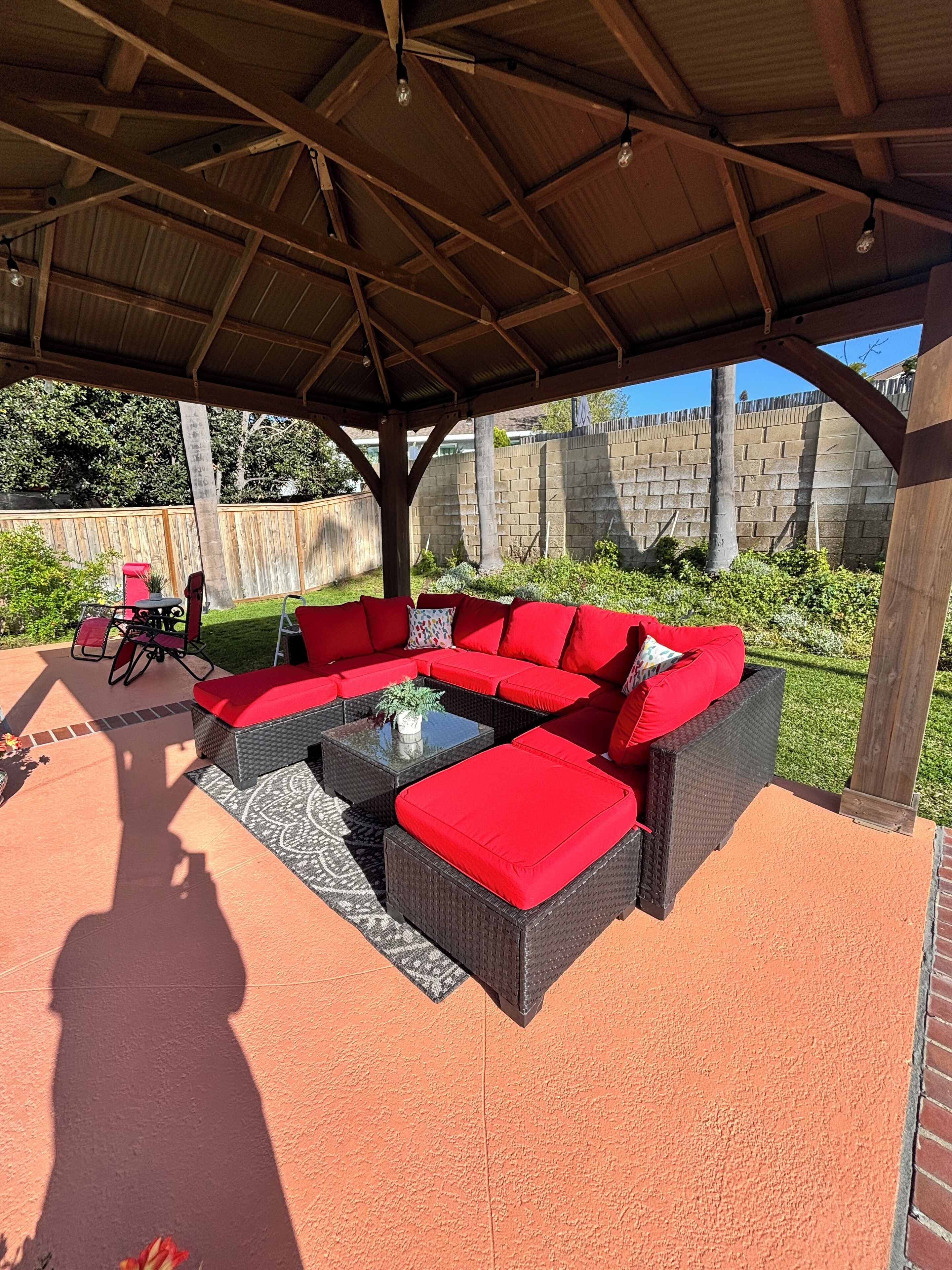 An outdoor patio with a sectional sofa and a dining table, surrounded by a wooden pergola and a brick wall.
