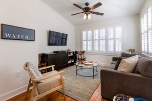 Uma sala de estar com um ventilador de teto, uma janela grande com persianas, uma televisão, uma estante de livros e uma placa com a inscrição 'WATER'.