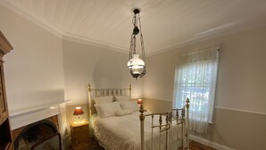 Un dormitorio con cama con dosel, una mesita de noche con lámpara, suelo de madera y una ventana con cortinas translúcidas.