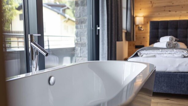 A modern bathroom with a freestanding bathtub, a wooden floor, and a bed with neatly folded towels.