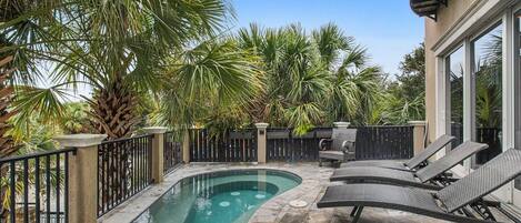 A swimming pool with a curved edge, surrounded by a stone deck and a metal fence. There are lounge chairs and a small patio table.