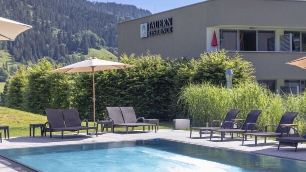 Zona de piscina con sillones reclinables y sombrillas, un edificio que dice "TAUERN RESIDENCE" y montañas al fondo.