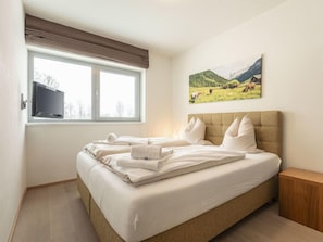 Un dormitorio con una cama grande, una ventana con vistas a un paisaje, un televisor montado y una mesita de noche de madera.