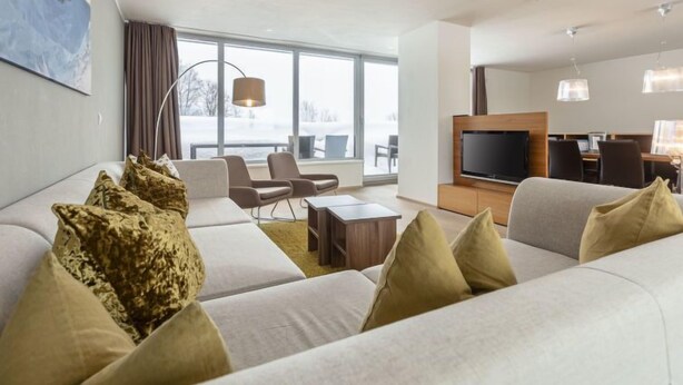 Una moderna camera d'albergo con un divano grande, una TV a schermo piatto, un'area pranzo e una grande finestra con vista su un paesaggio innevato.