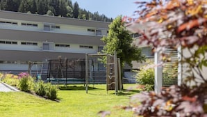 A grassy outdoor area with a playground, including a swing set and climbing structure, in front of a multi-story building with a flat roof.