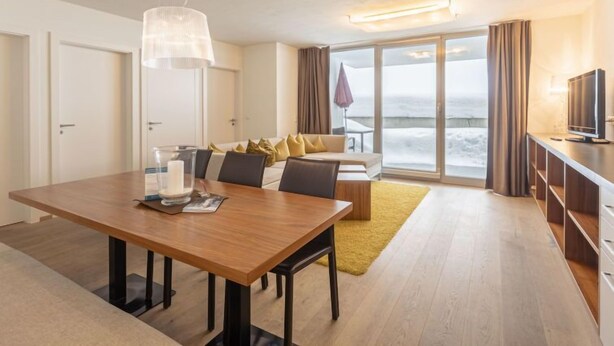 A modern hotel room with a wooden dining table, a sofa, a TV, and a large window with a view of the sea.