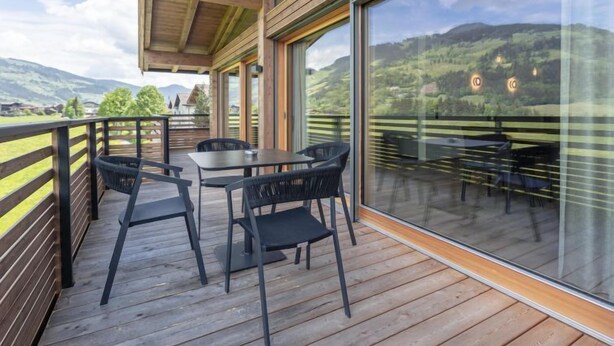 Een houten terras met een tafel en stoelen, grote glasramen en uitzicht op een bergachtig landschap.